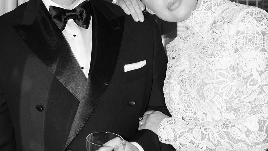 Man in a tuxedo holding a glass next to a woman in a white lace dress.